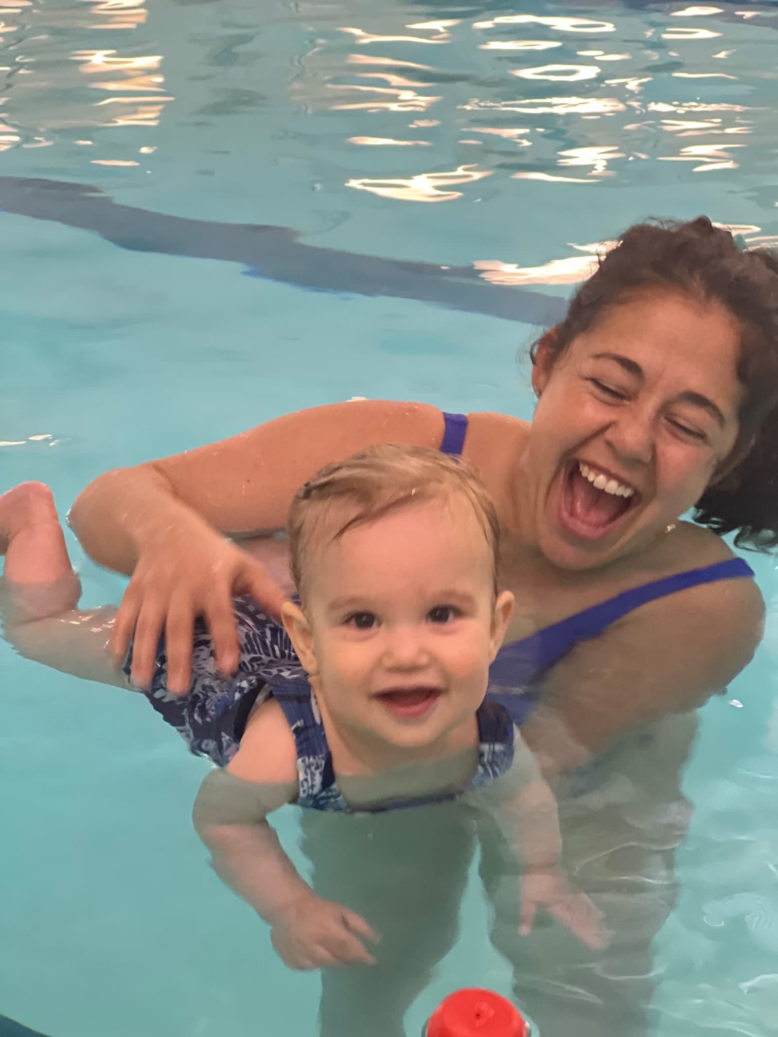 Joyful woman laughing while supporting a smiling baby in a bright blue swimming pool.